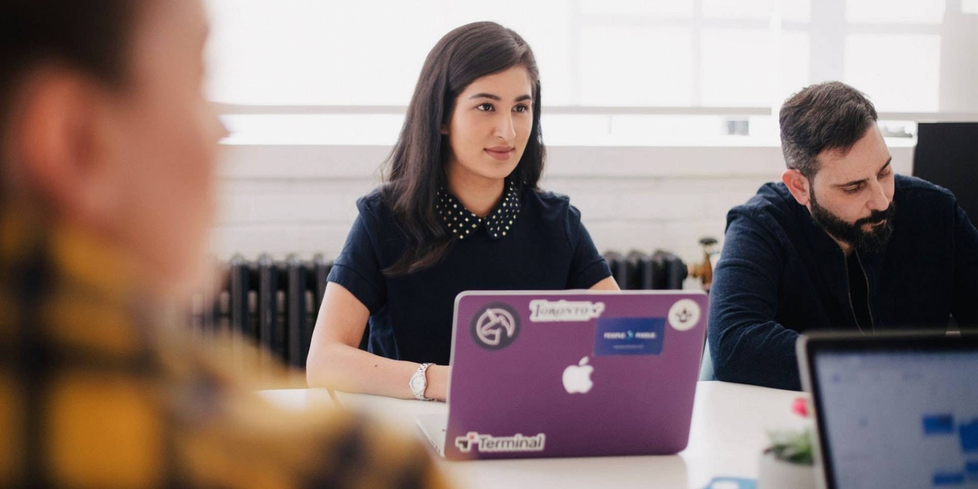 Two people in meeting, a woman paying attention from behind a MacBook.