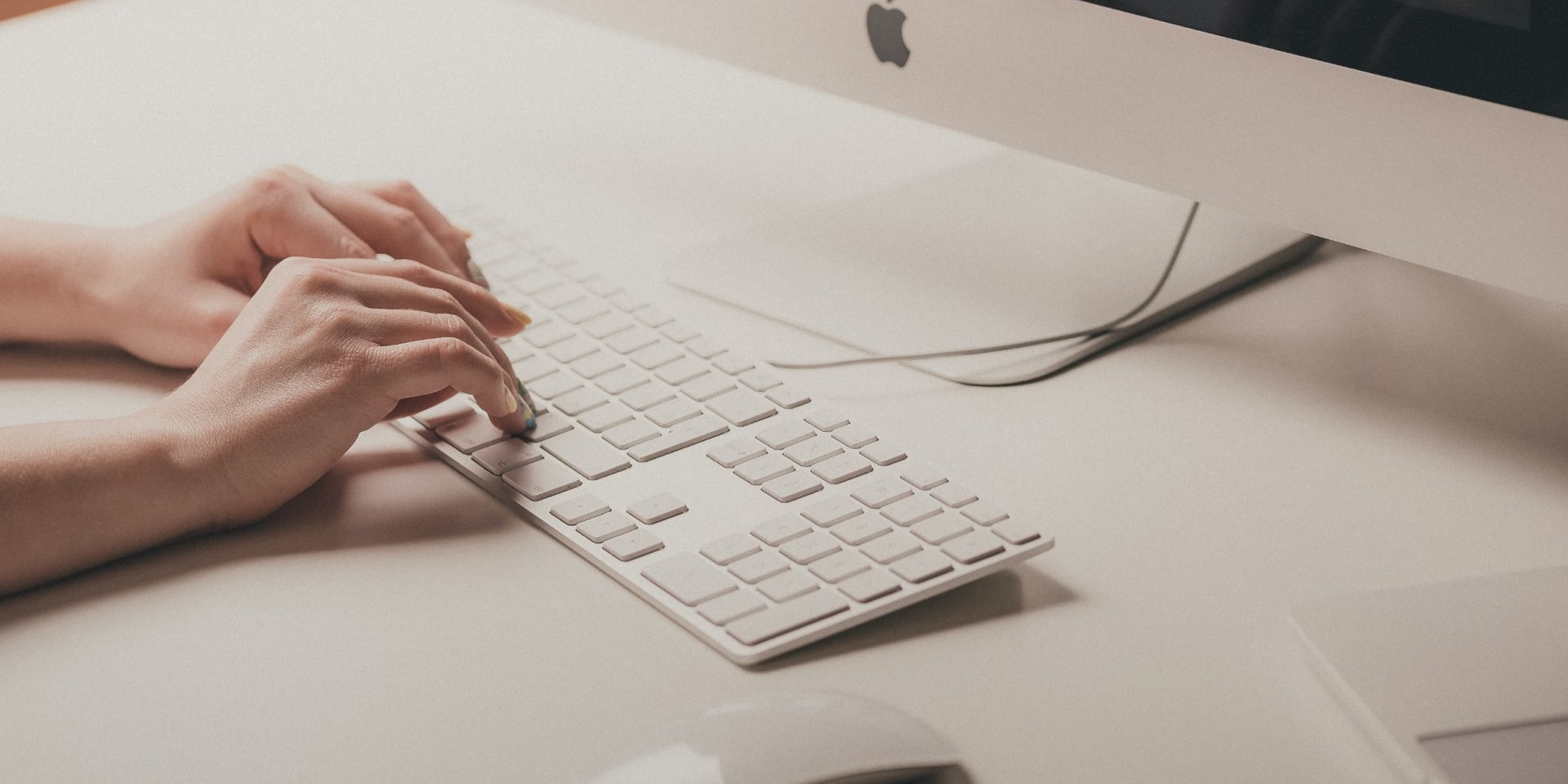 Two hands typing behind a keyboard on desk where a mouse and monitor is slightly visible.